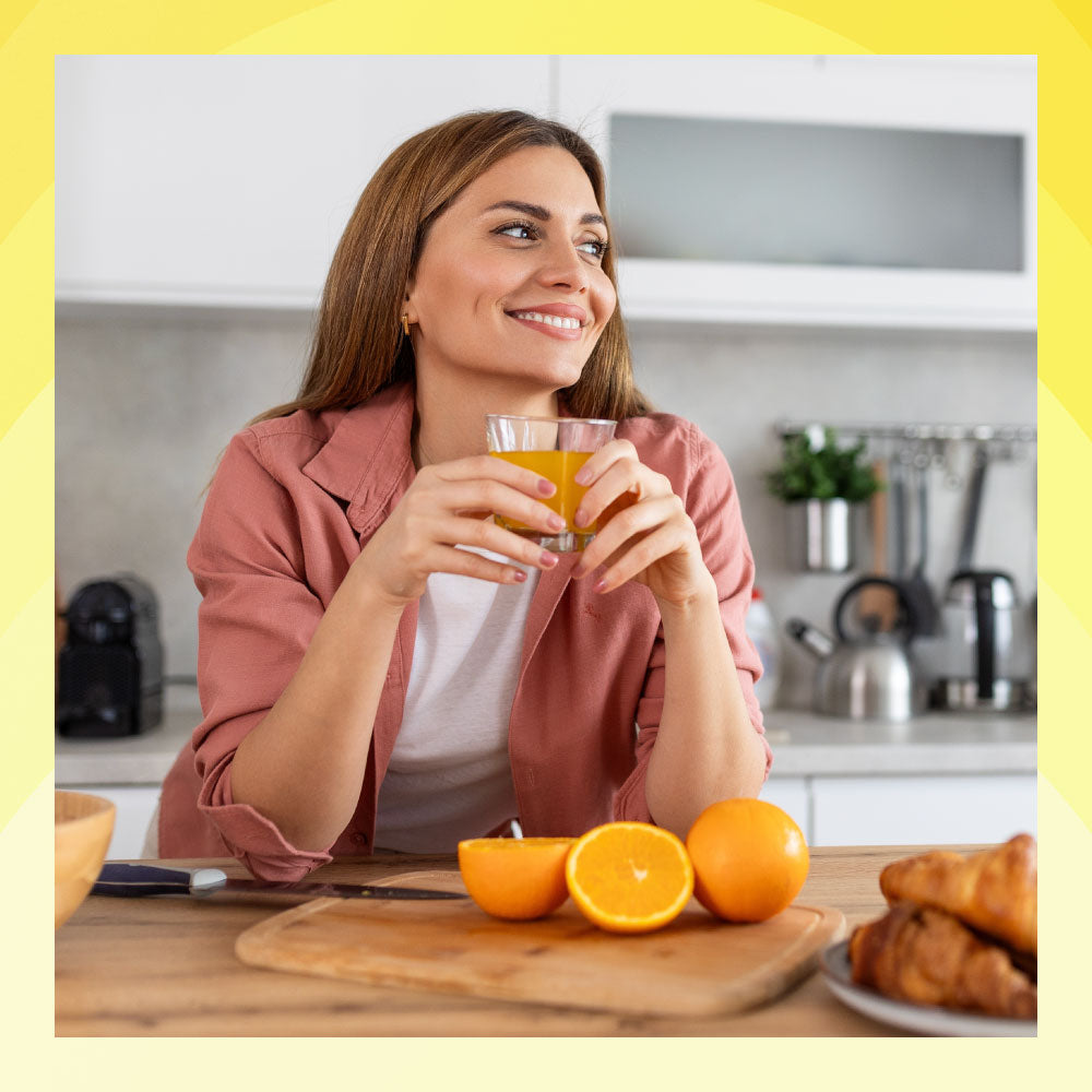 Woman holding a glass of orange juice in a kitchen with oranges and pastries on the table.